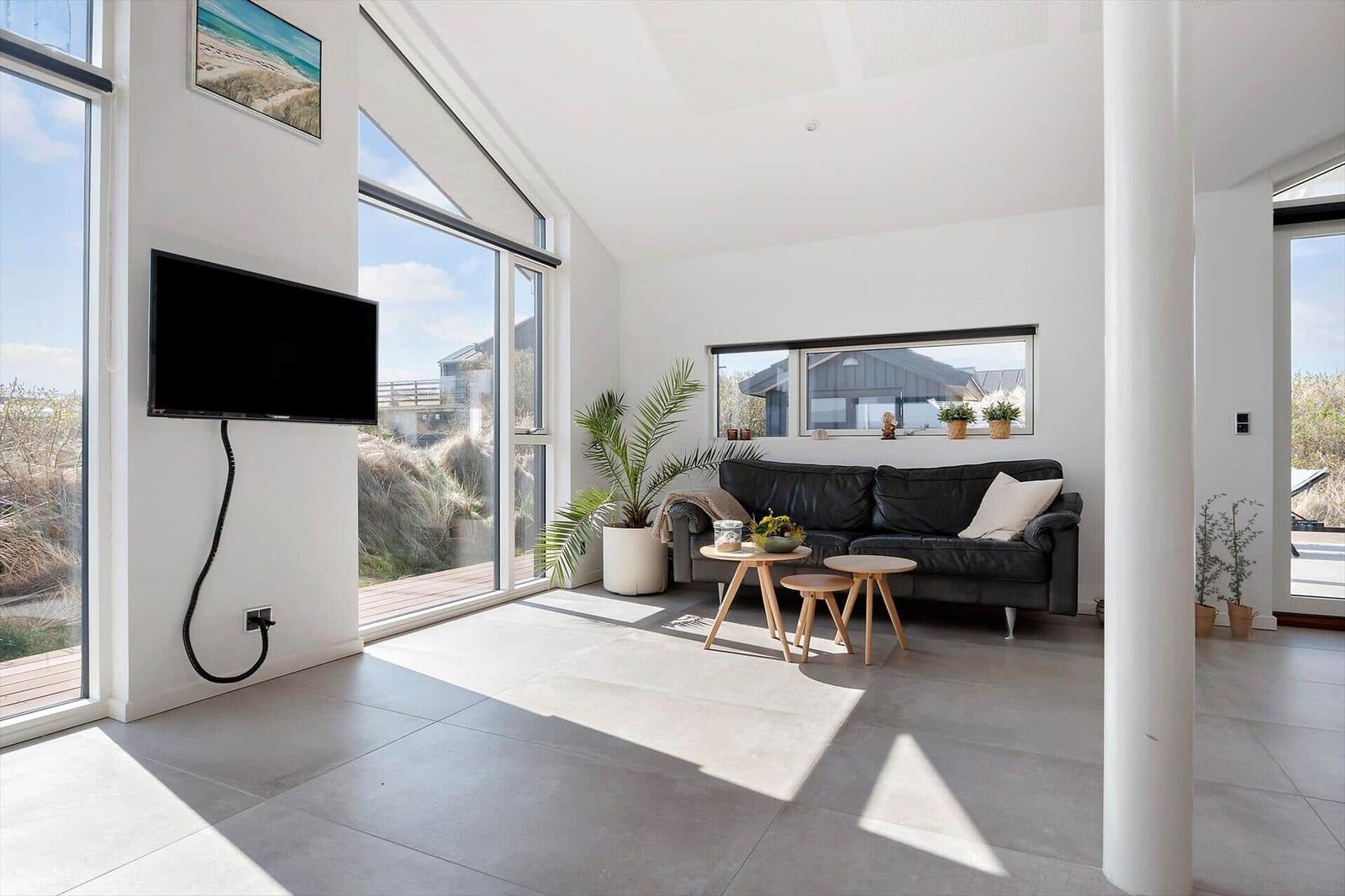 Wohnzimmer mit schwarzer Couch, TV und großen Fenstern. Draußen ist eine Terrasse.