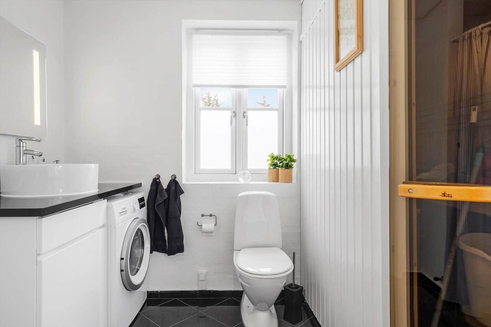 Badezimmer mit Waschmaschine, Toilette und Fenster mit Pflanzen.