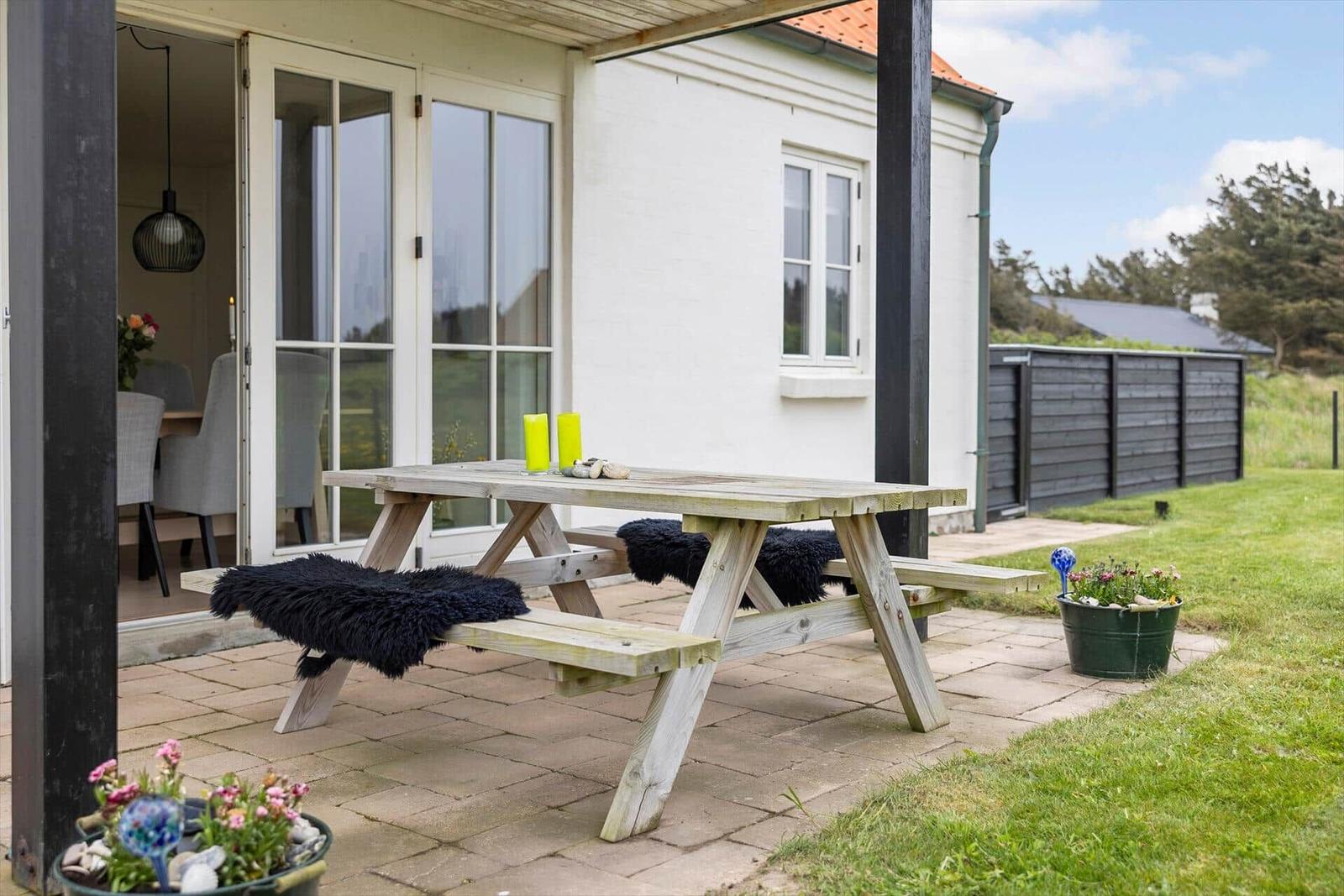 Holzpicknicktisch mit Felldecken auf Terrasse neben weißem Haus mit Glastüren.