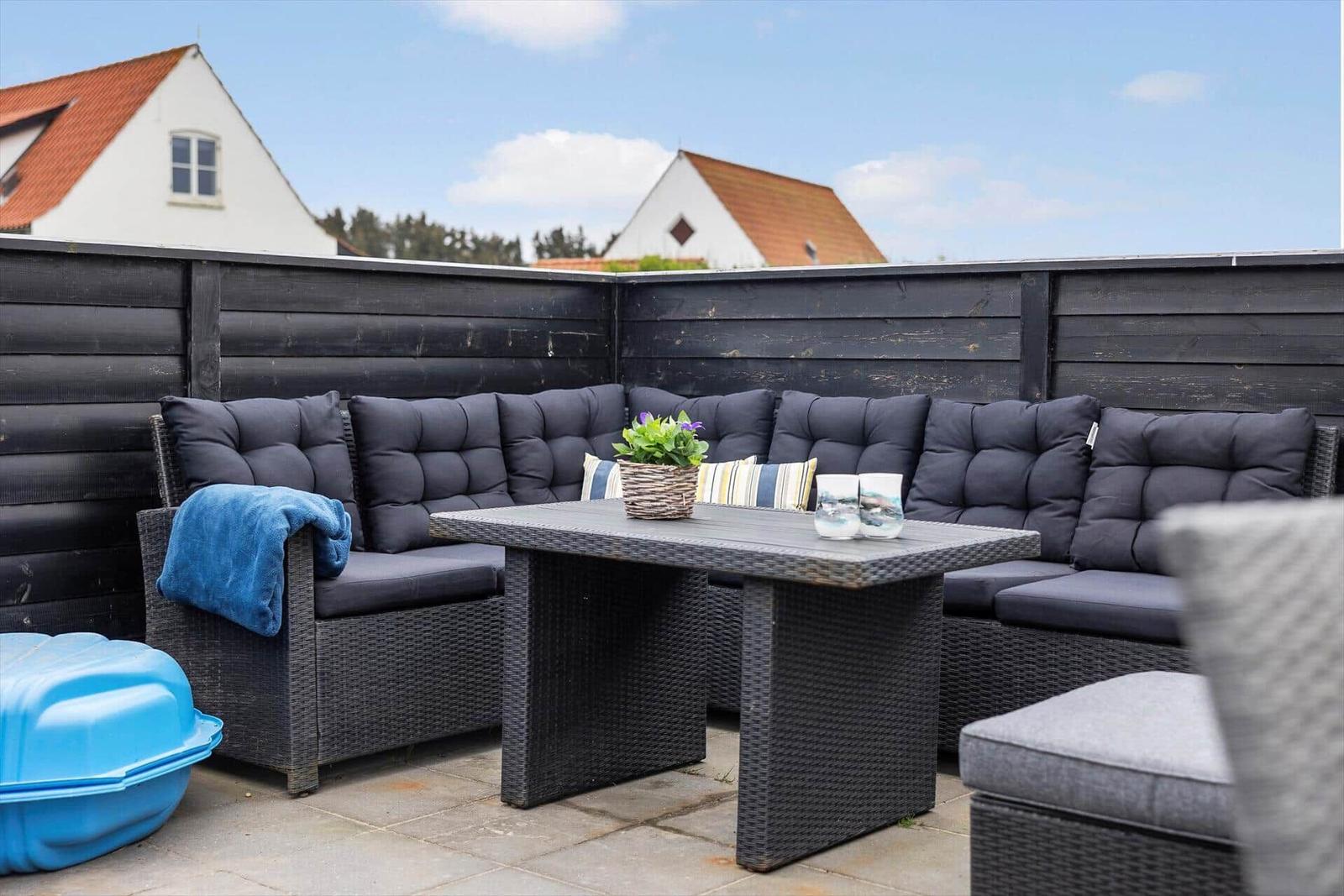 Terrasse mit Sitzgruppe, Tisch und Pflanze. Hintergrund: Holzzaun und Häuser.