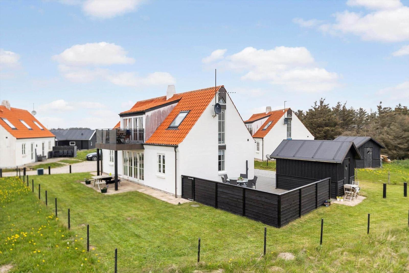 Wohnhaus mit rotem Dach, Terrasse und Garten. Schwarze Holzumzäunung und Nebengebäude.