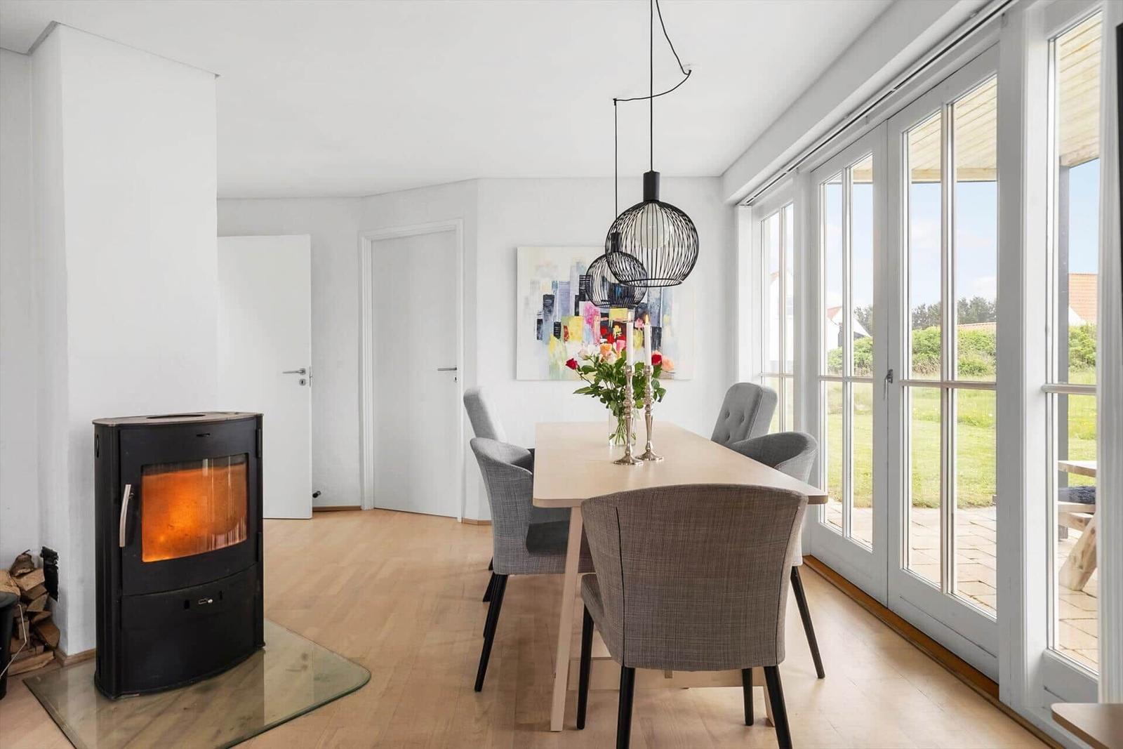 Esszimmer mit Holztisch, Stühlen und Kaminofen. Große Fensterfront mit Blick auf Garten.