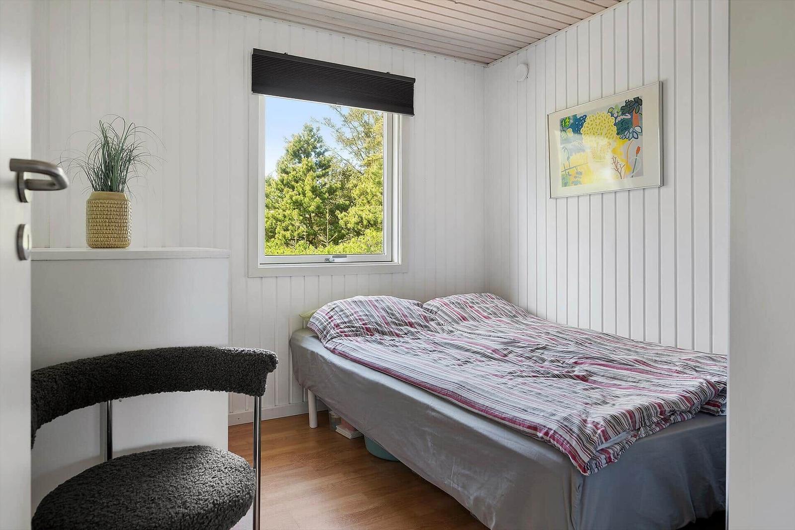 Schlafzimmer mit Bett, Fenster und Stuhl. Wand mit Bild und Pflanze.