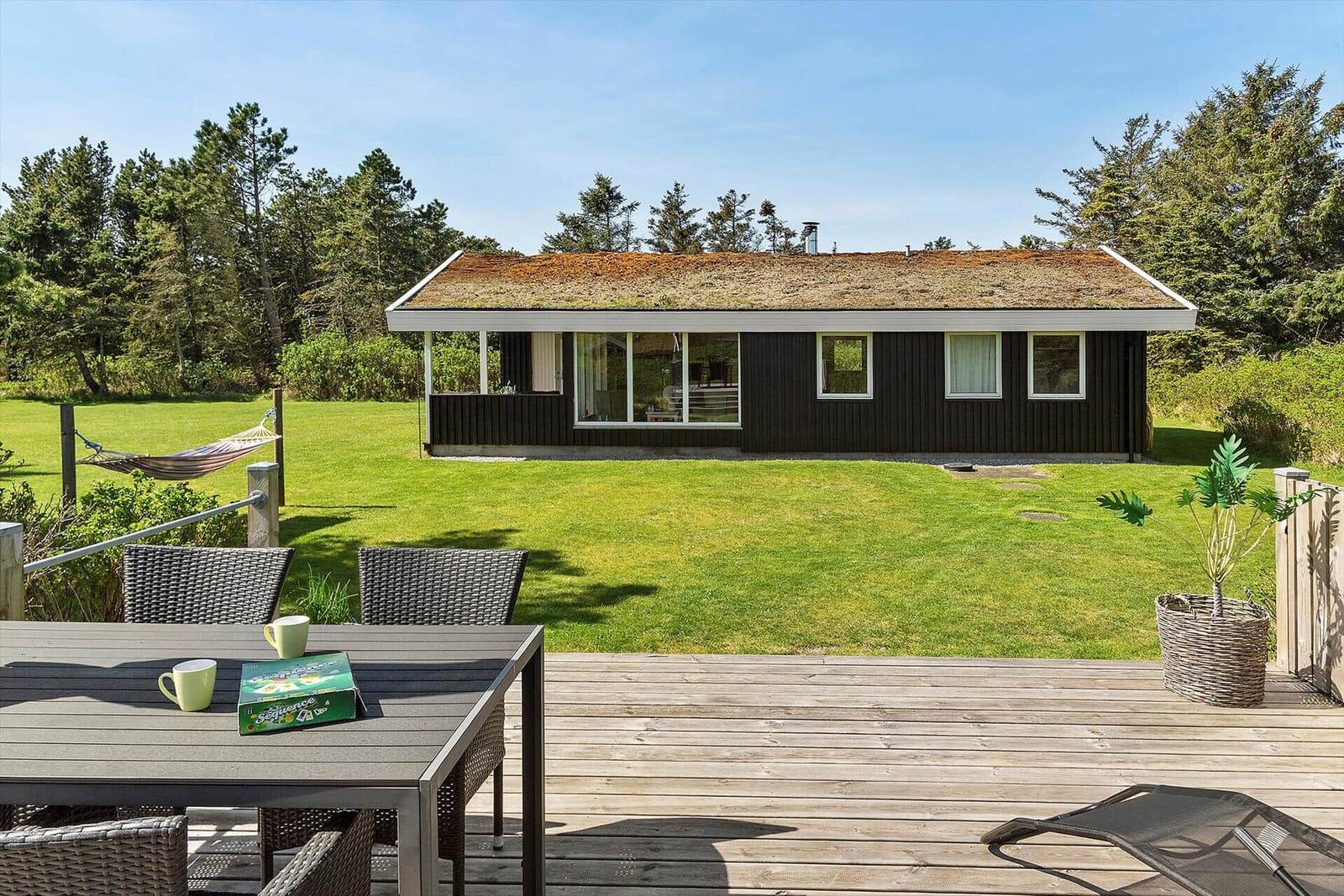 Holzterrasse mit Tisch, Stühlen und Hängematte vor einem Haus mit Grasfläche und Bäumen.