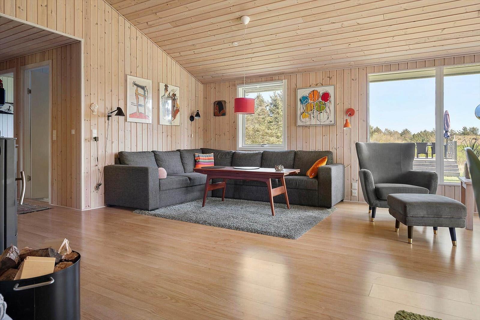 Wohnzimmer mit Holzwand, grauem Sofa, Sessel und großem Fenster.