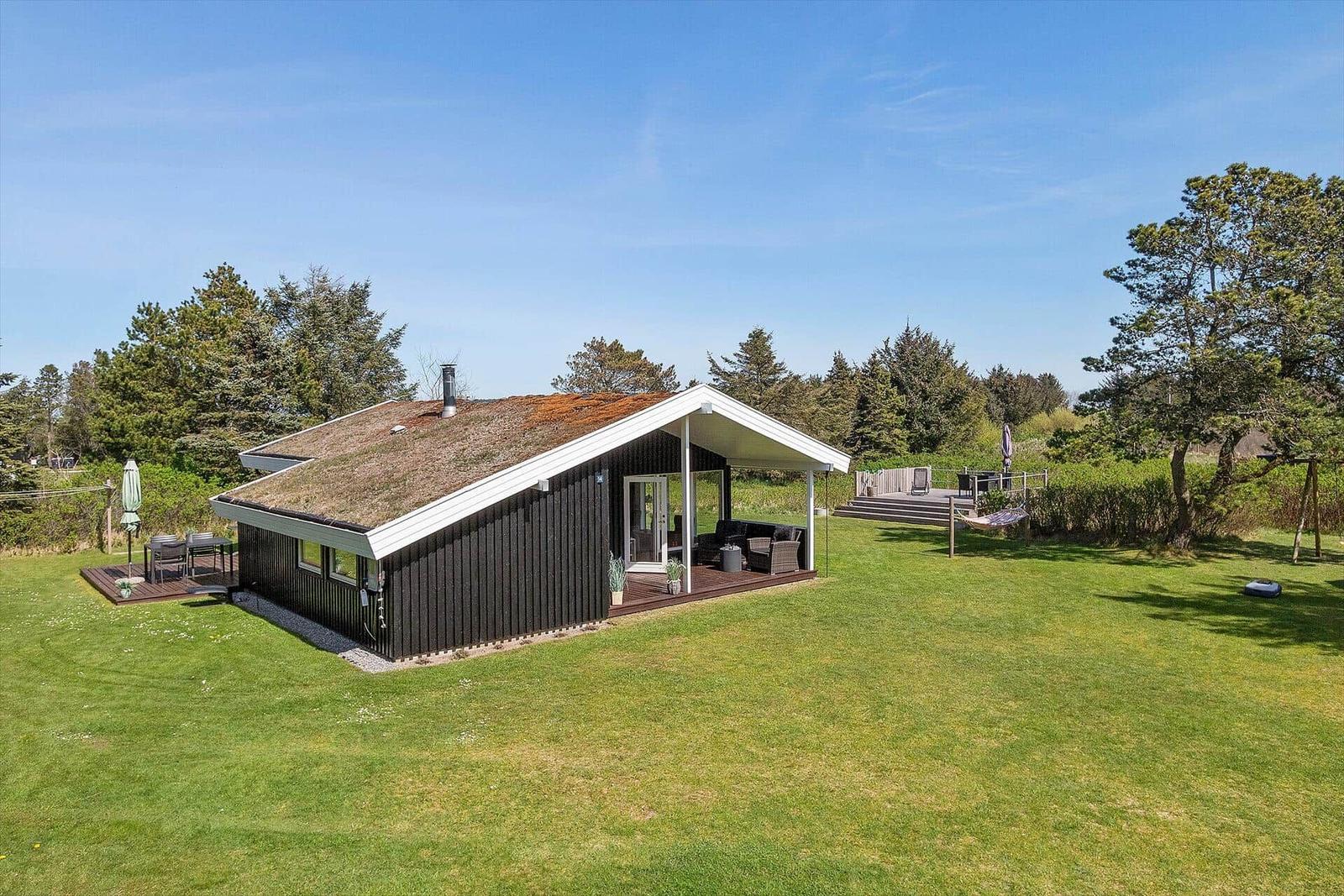 Haus mit Grasdach, Terrasse und Garten. Umgeben von Bäumen und Grasflächen.