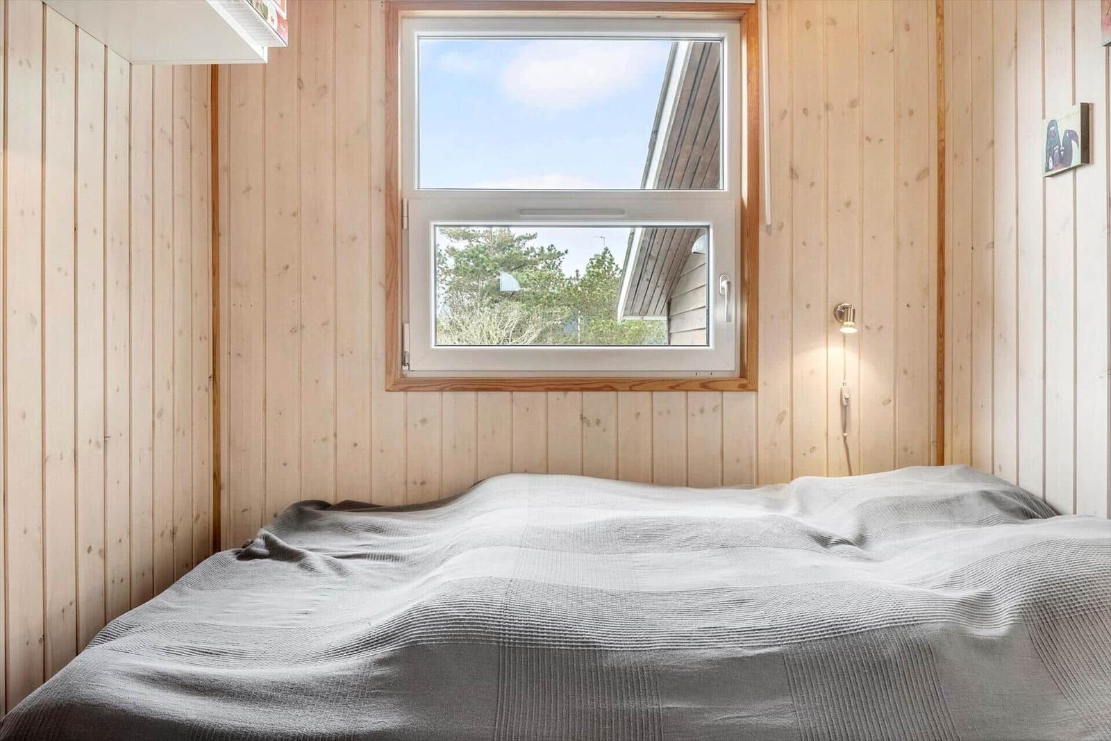 Ein Schlafzimmer mit Holzwänden, einem Fenster und einem Bett mit grauer Bettwäsche.