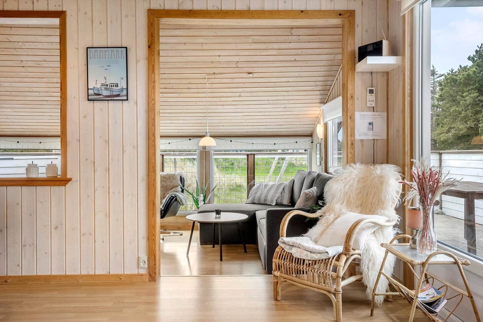 Wohnraum mit Holzwänden, Sofa und Sessel. Fenster zeigen Grünfläche. Wandbild zeigt Boot.