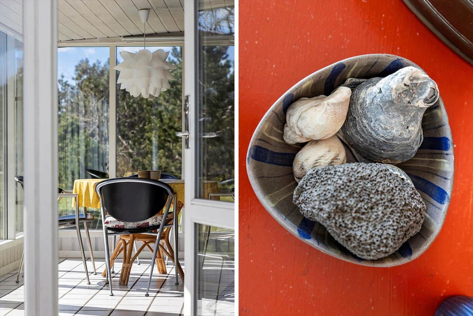 Terrasse mit Tisch und Stühlen, Blick auf Bäume. Schale mit Muscheln und Steinen auf roter Wand.