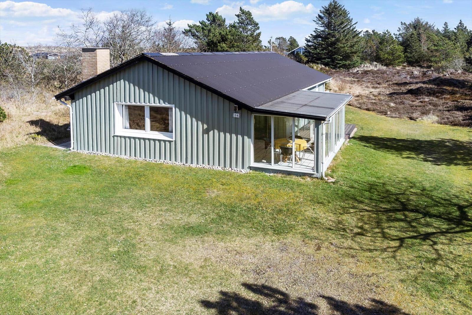Ein graues Haus mit Glasveranda und Garten. Im Inneren ist ein Tisch mit gelben Stühlen zu sehen.