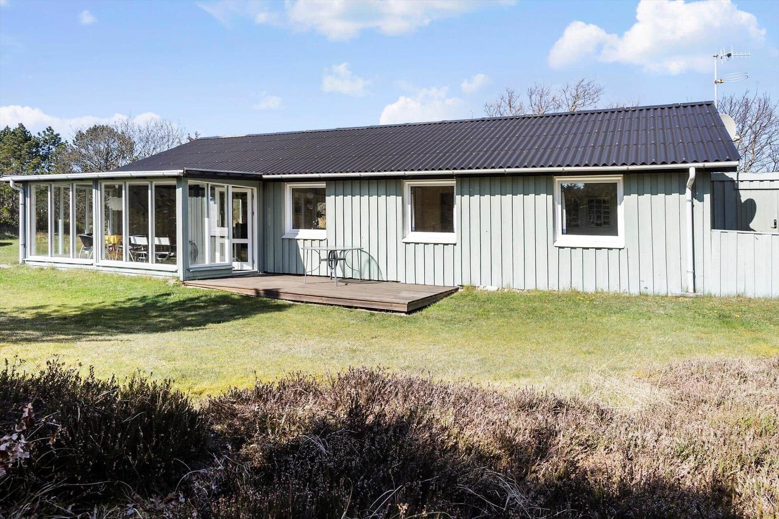Ein Haus mit Holzfassade, Terrasse und Glasfront. Grünfläche und Sträucher im Vordergrund.