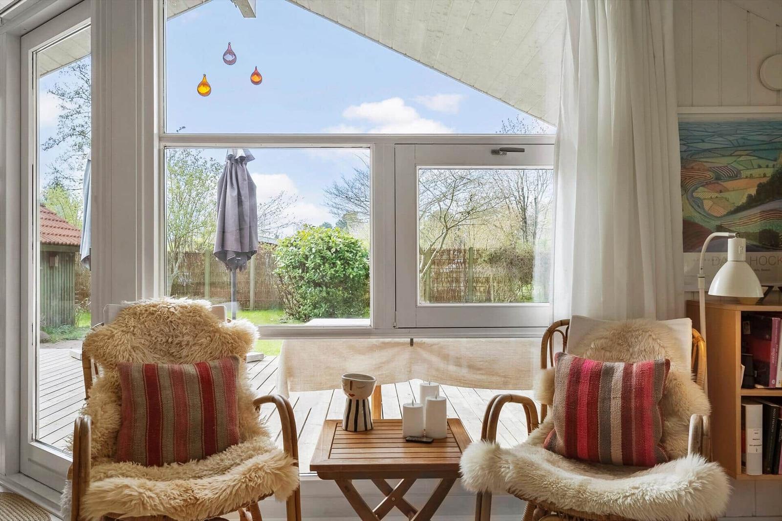 Zwei Stühle mit Fell und Kissen stehen vor einem großen Fenster mit Blick auf eine Terrasse.