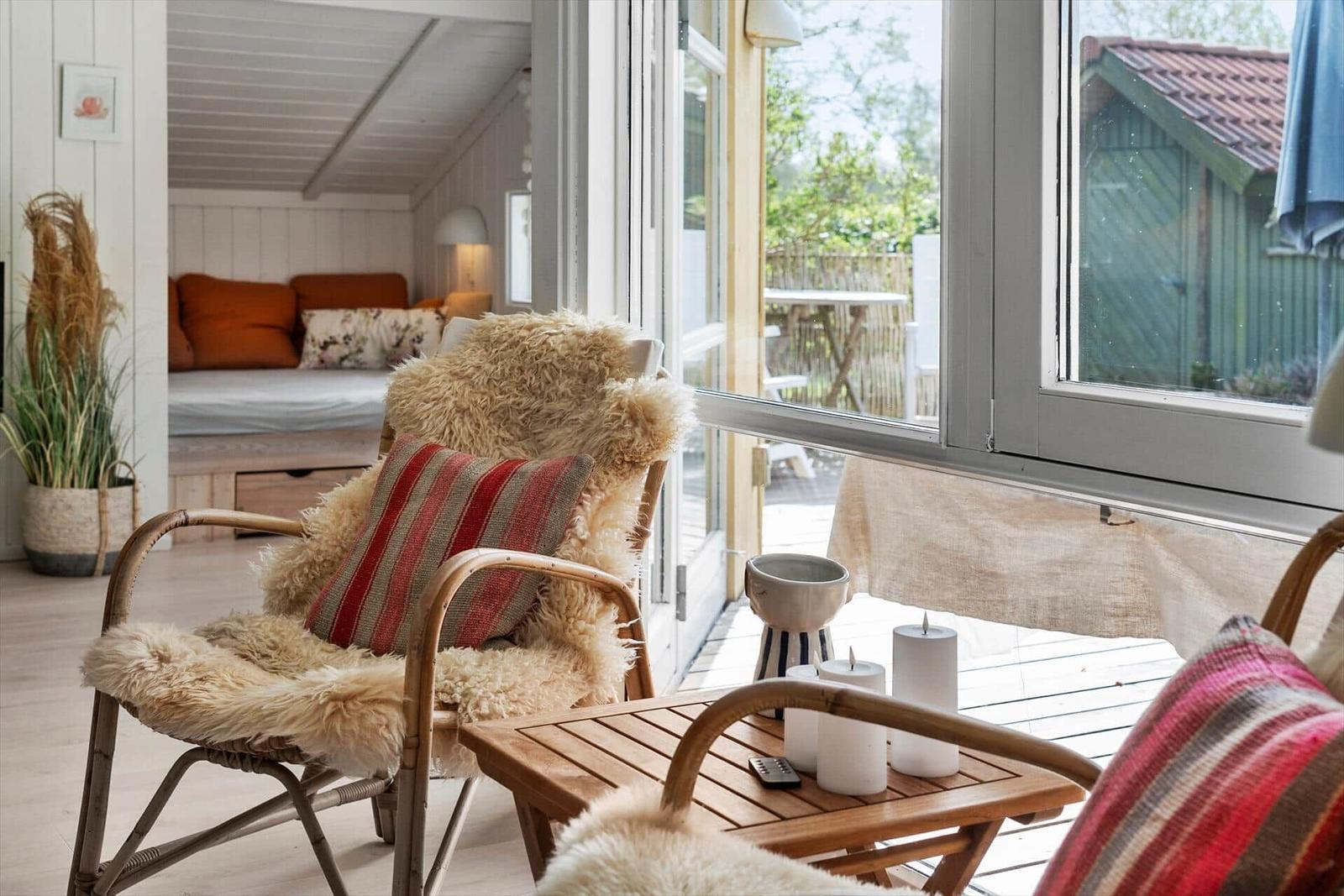 Sitzgelegenheit mit Felldecke und Kissen vor großem Fenster. Im Hintergrund Schlafbereich mit orangefarbenen Kissen.