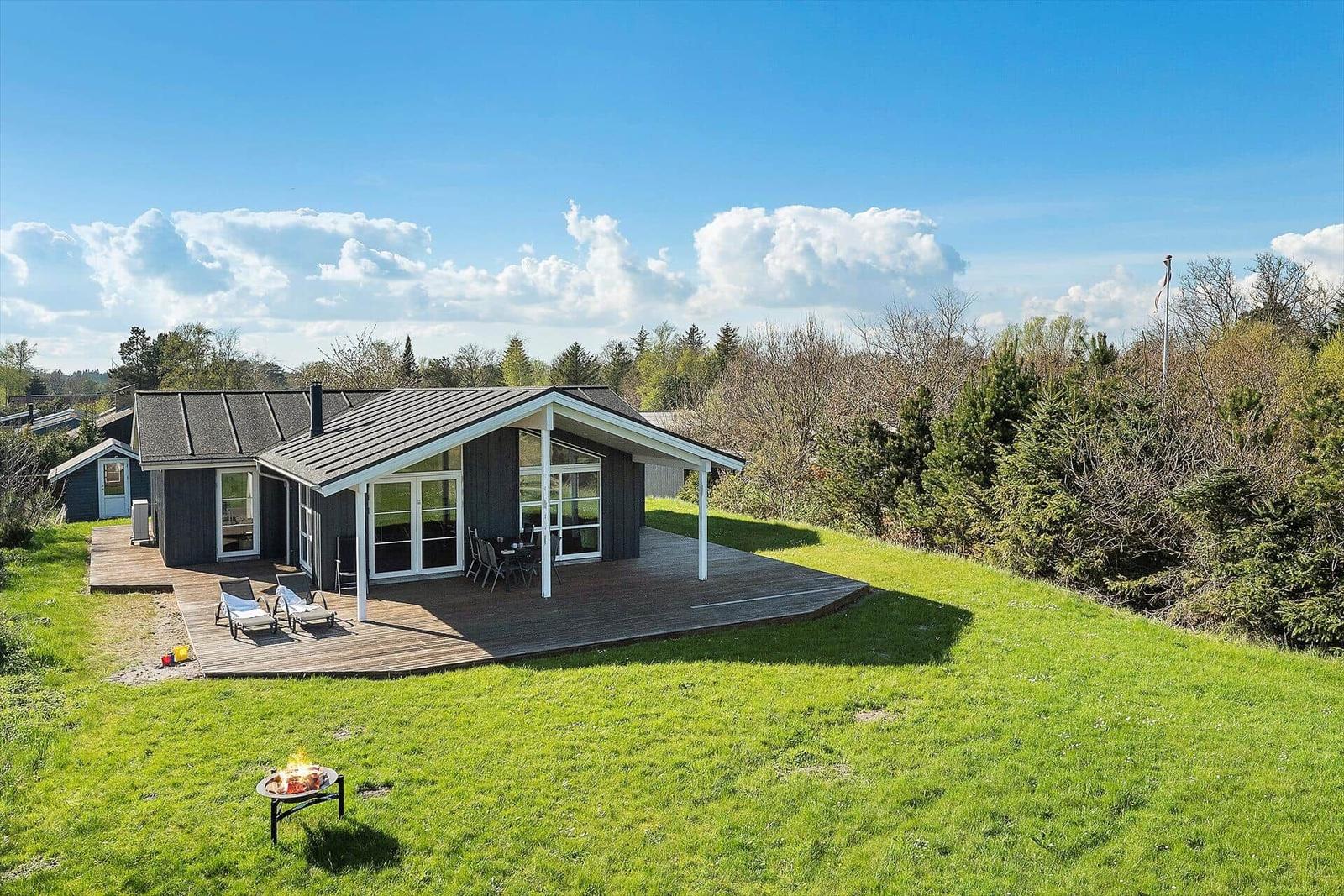 Moderne Hütte mit Terrasse, Garten und Feuerkorb unter blauem Himmel.
