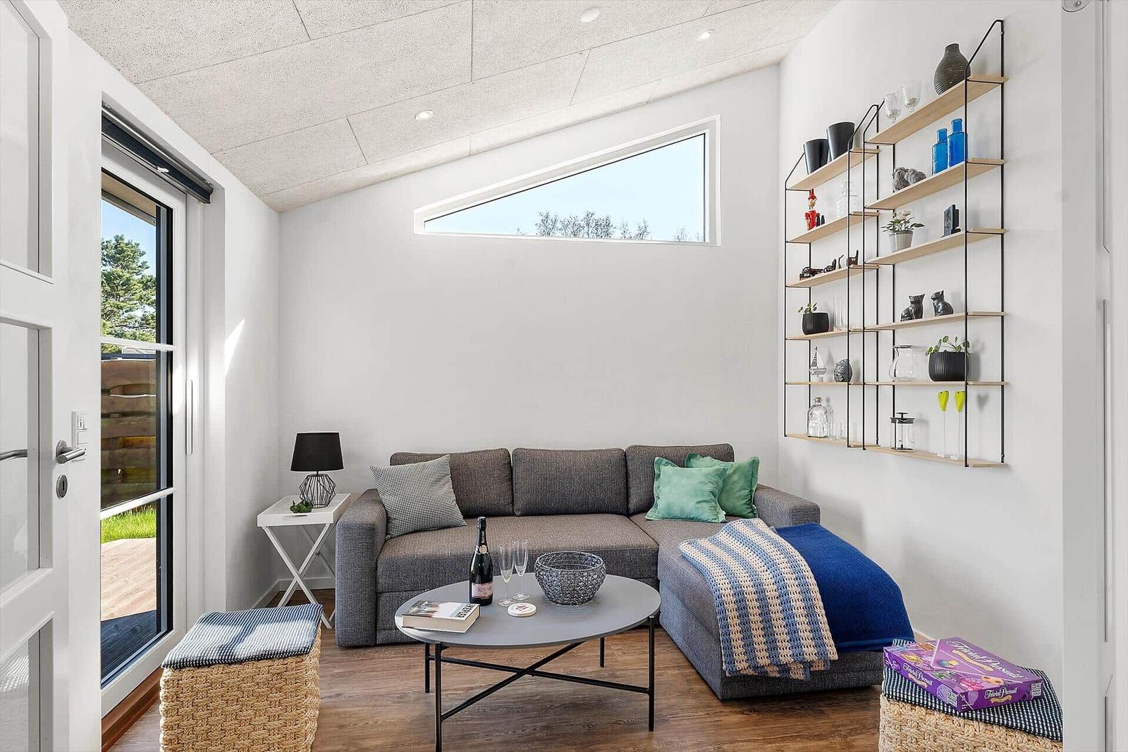 Wohnzimmer mit Sofa, Couchtisch und Wandregal. Fenster und Tür führen nach draußen.