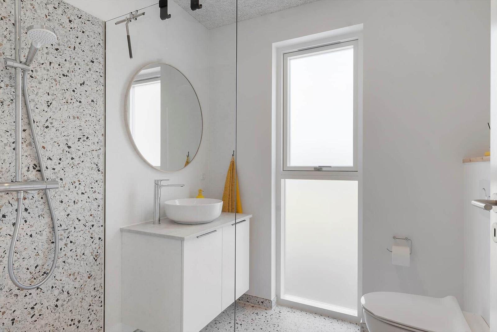 Badezimmer mit Dusche, Waschbecken und Toilette. Terrazzo-Wand und Fenster.