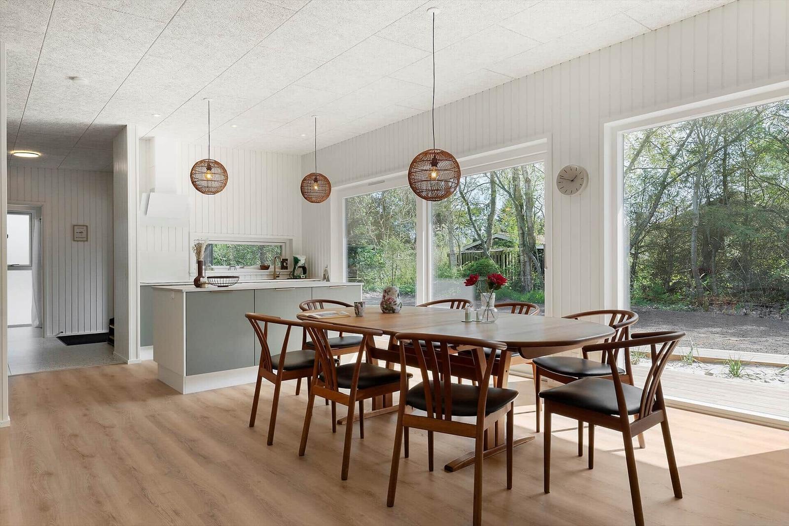 Essbereich mit Holztisch, Stühlen und Küchenzeile. Große Fenster zeigen Wald.