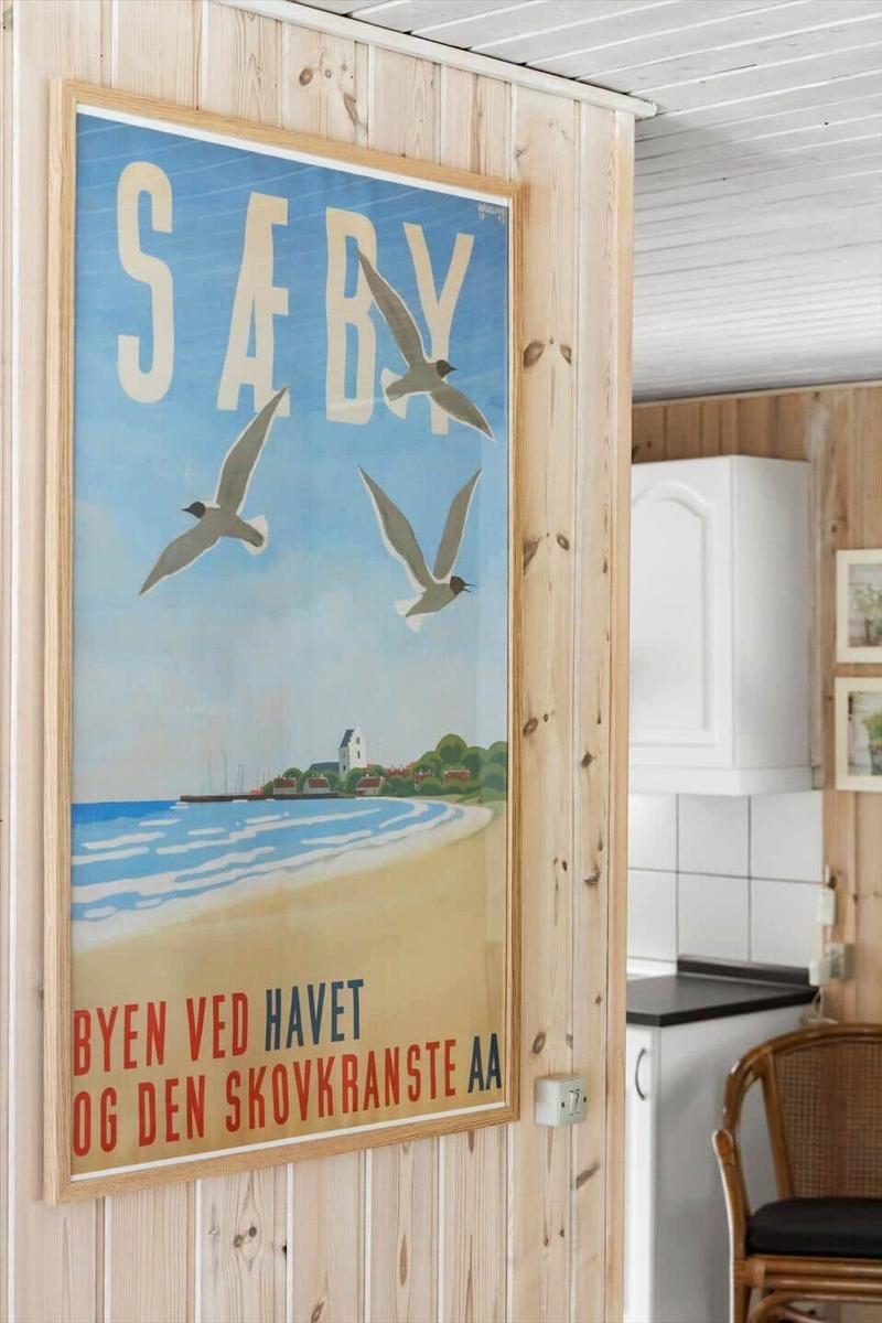 Wand mit Poster von Säby, Küchenbereich mit Holzwand und Stuhl.