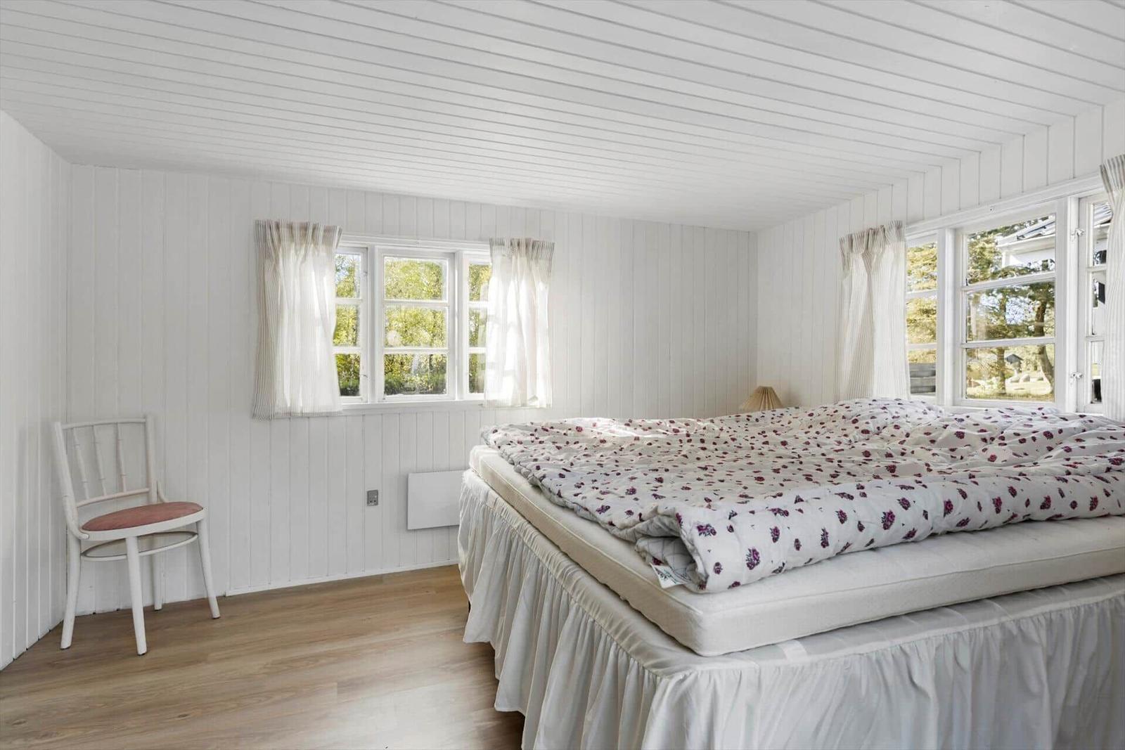 Schlafzimmer mit Bett, Fenstern und Stuhl. Weiße Wände und Holzboden.