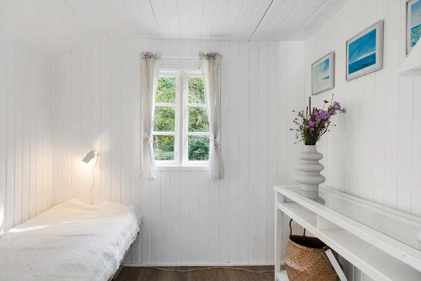 Ein Zimmer mit weißem Holzverkleidung, Fenster, Bett und Vase mit Blumen.