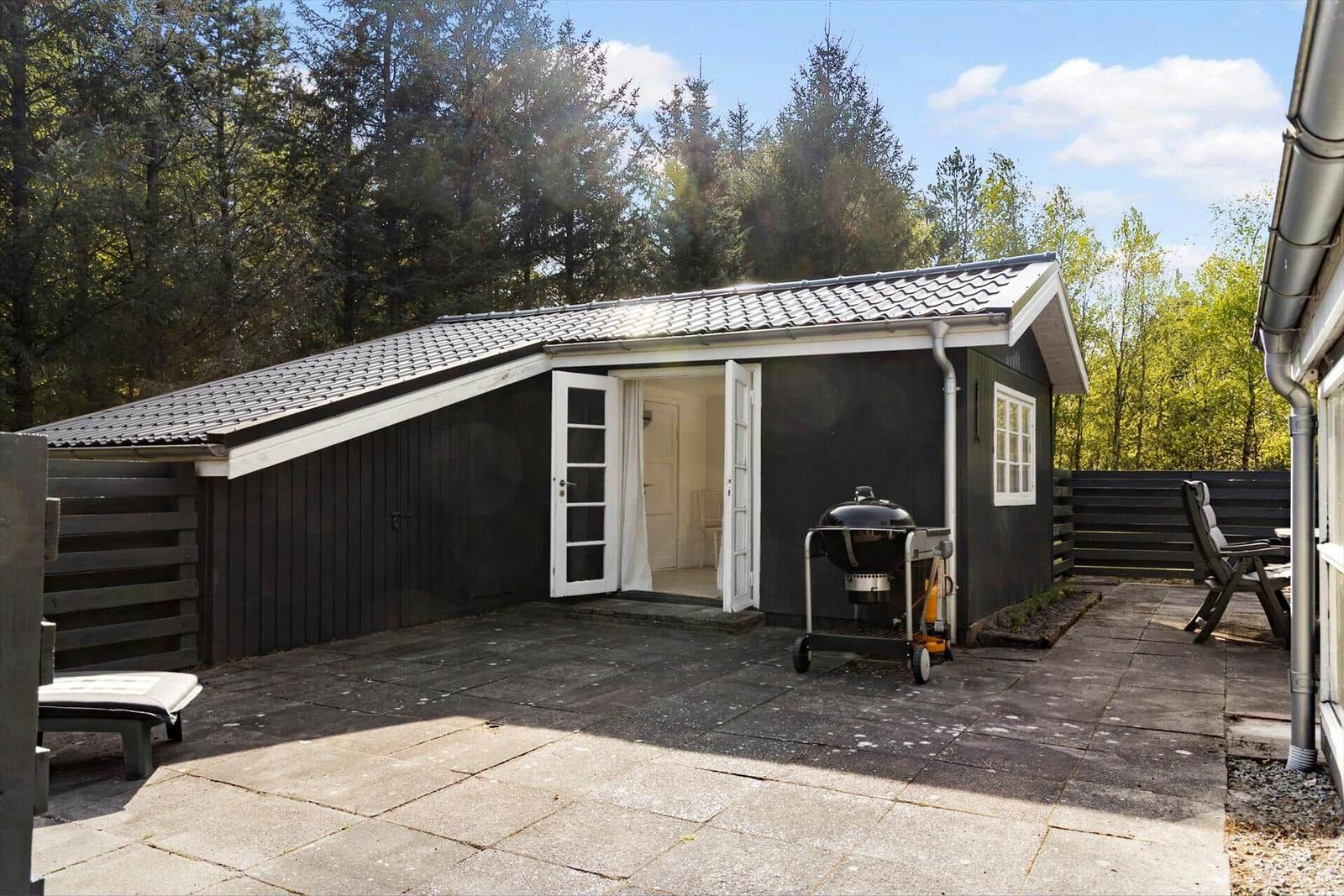 Ein kleines Haus mit offener Tür, Grill und Terrasse im Wald.