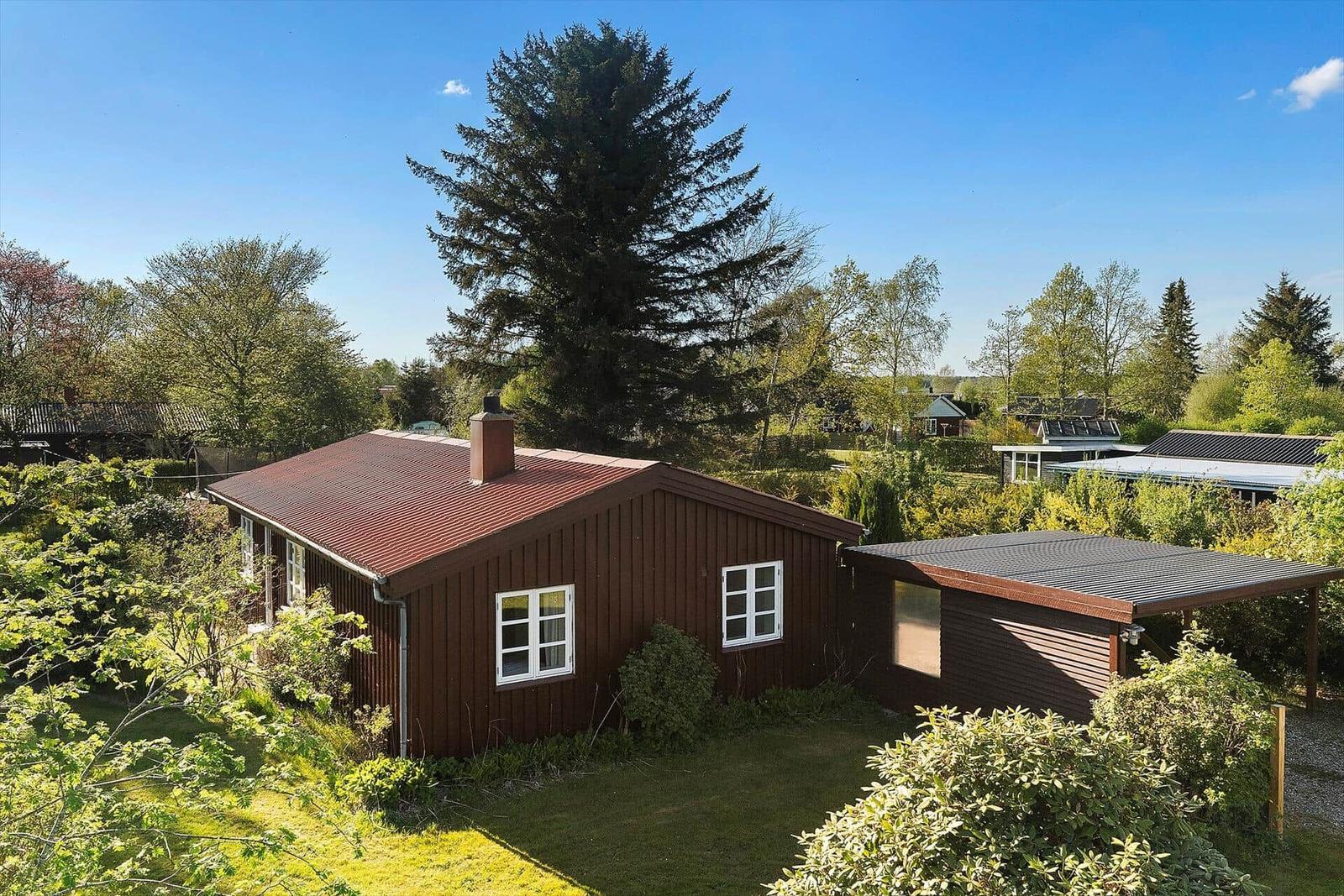 Ein braunes Holzhaus mit rotem Dach und weiß gerahmten Fenstern inmitten grüner Bäume und Sträucher.
