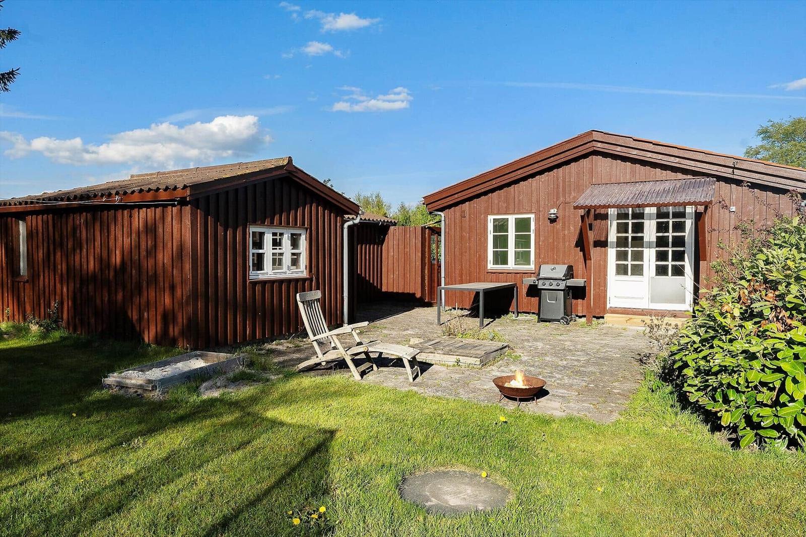 Holzhütte mit Grill und Feuerkorb im Garten unter blauem Himmel.