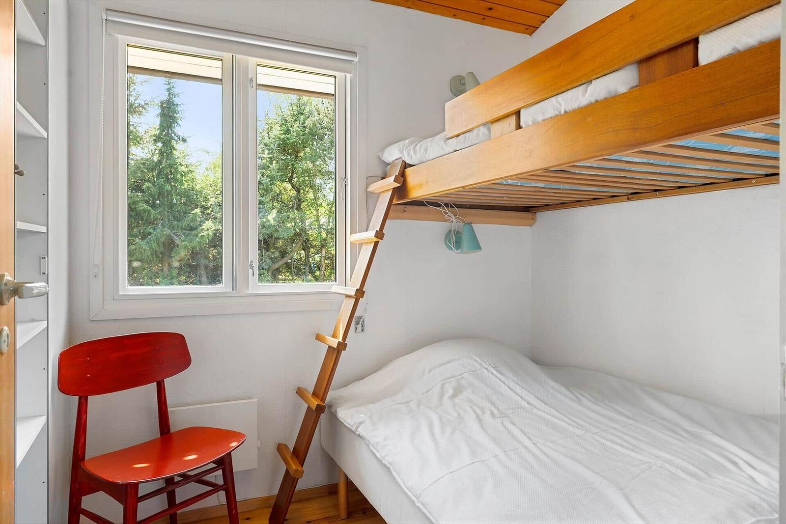 Ein Schlafzimmer mit Doppelstockbett, roter Stuhl und Fensterblick auf Bäume.