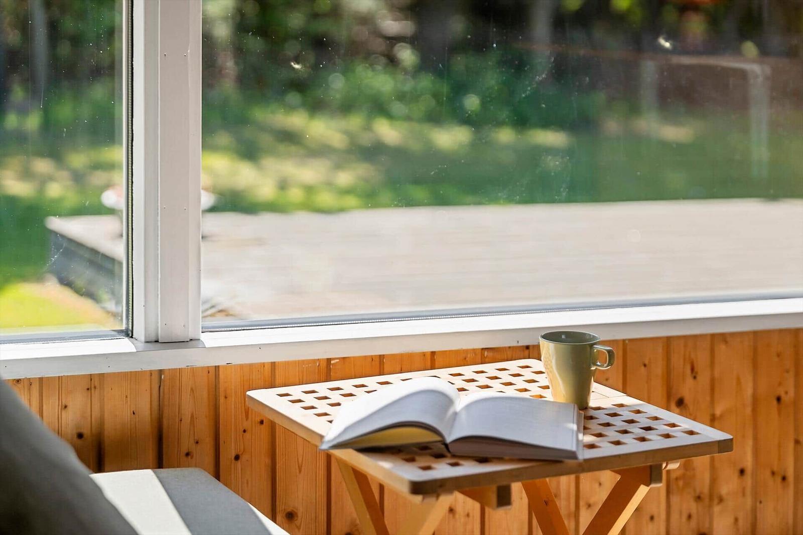 Ein Tisch mit Buch und Tasse vor einem Fenster mit Blick auf den Garten.
