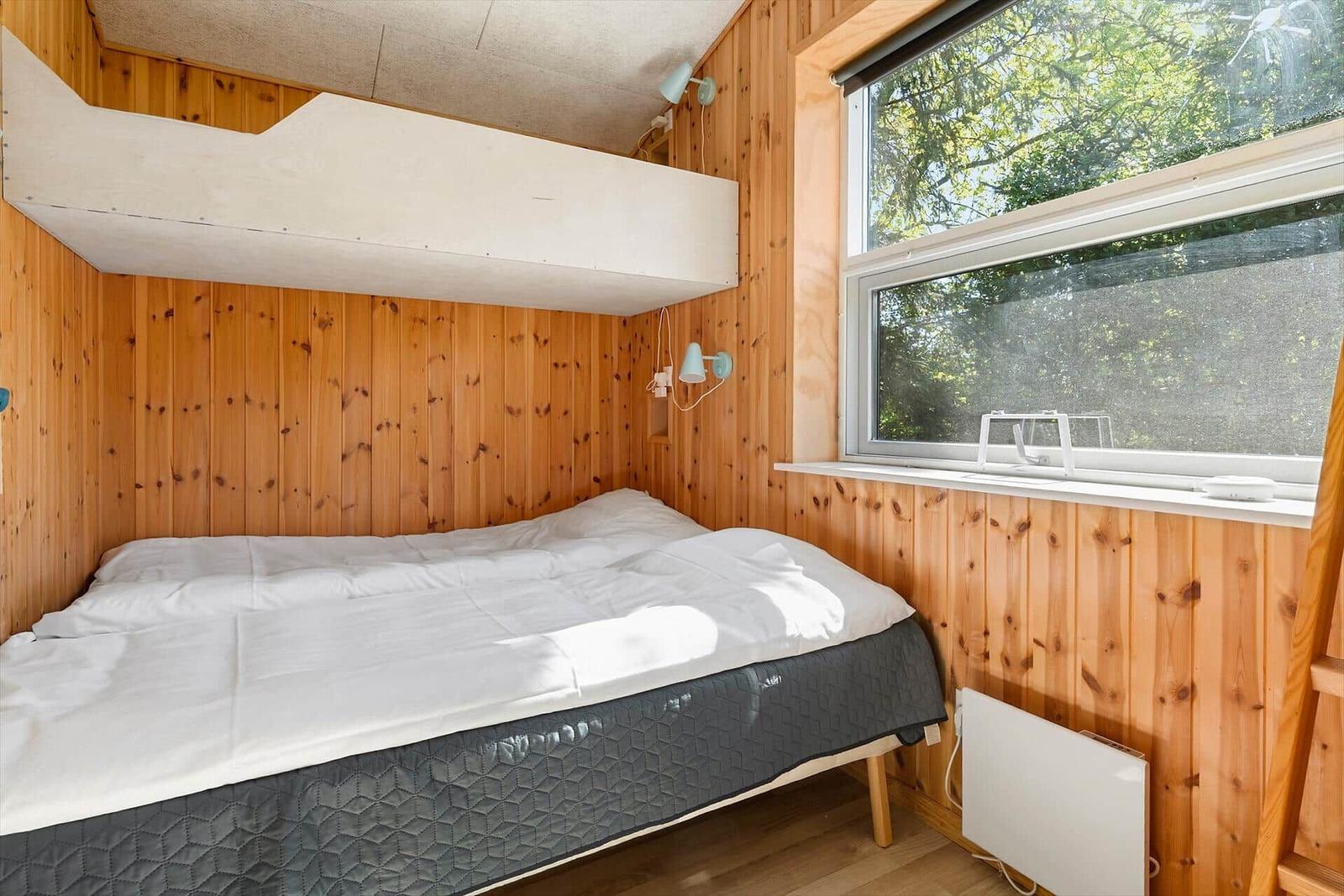 Schlafzimmer mit Bett, Holzwand und Fenster mit Blick auf Bäume.