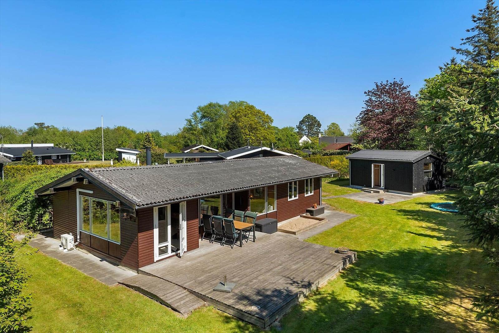 Ein Ferienhaus mit Holzterrasse, Garten und Pool. Umgeben von Bäumen und Grünflächen.