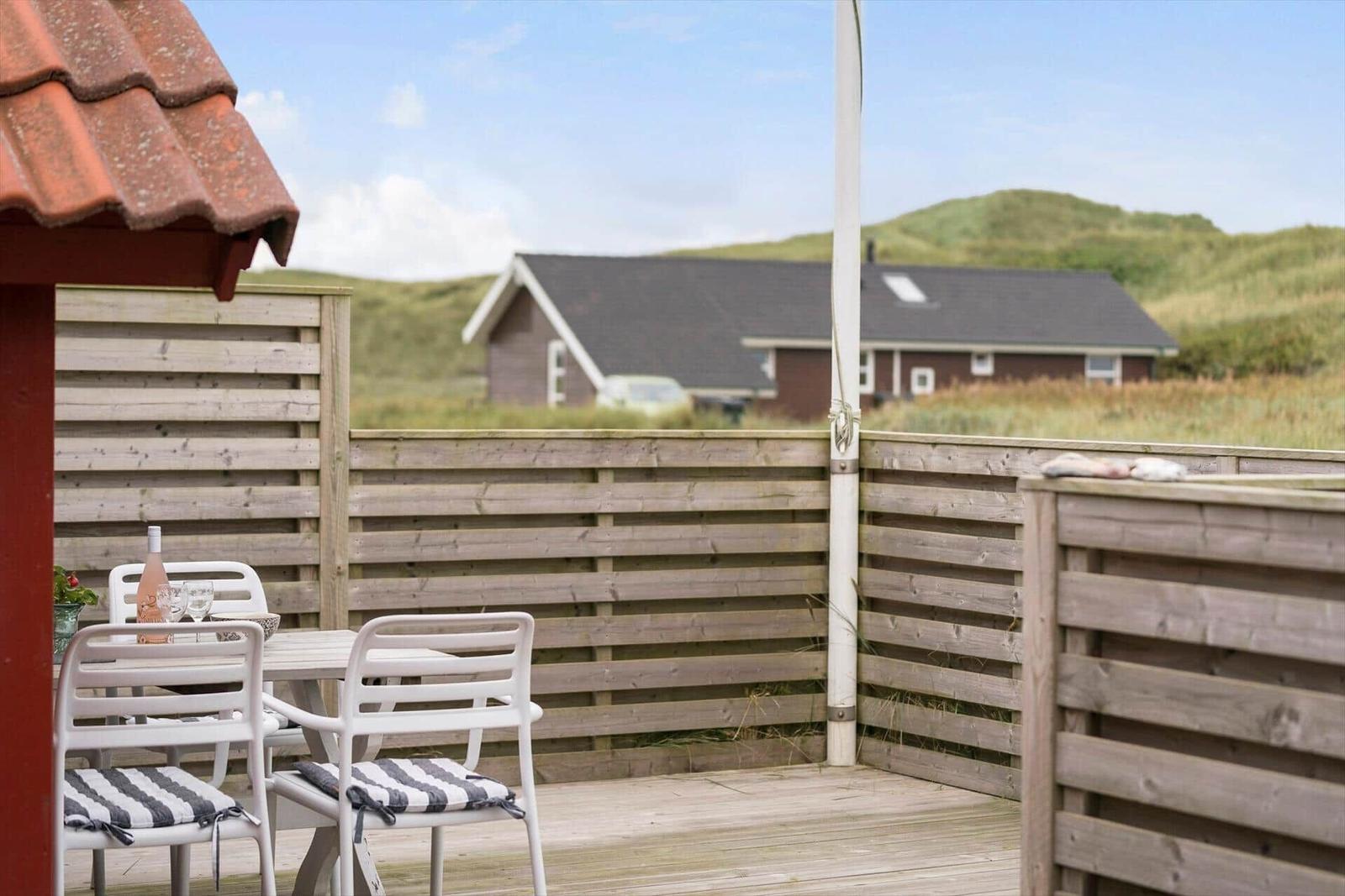 Terrasse mit Tisch und Stühlen, Holzzaun, Blick auf grüne Hügel und Haus im Hintergrund.