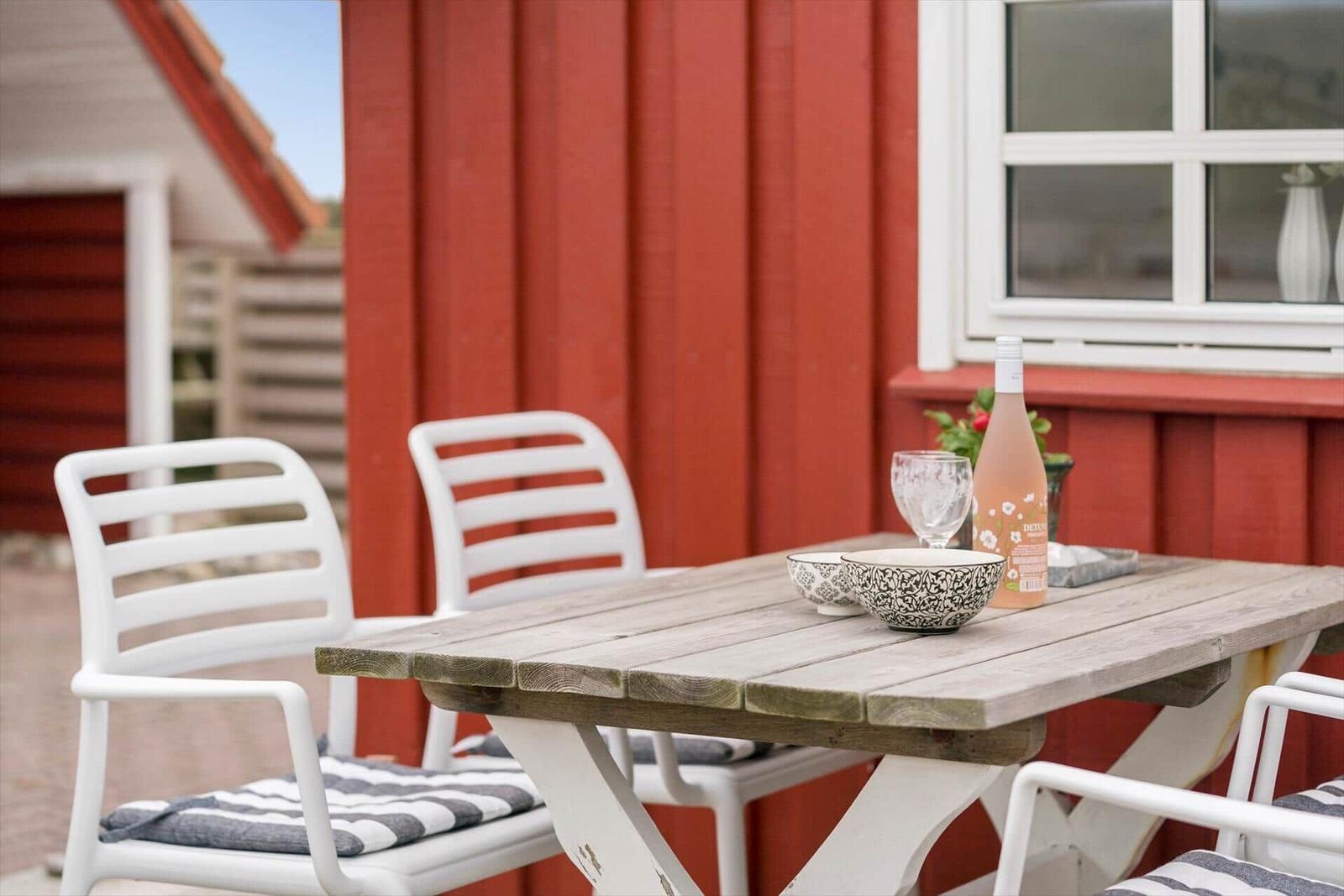 Holztisch mit Stühlen vor rotem Haus mit Flasche und Schalen.