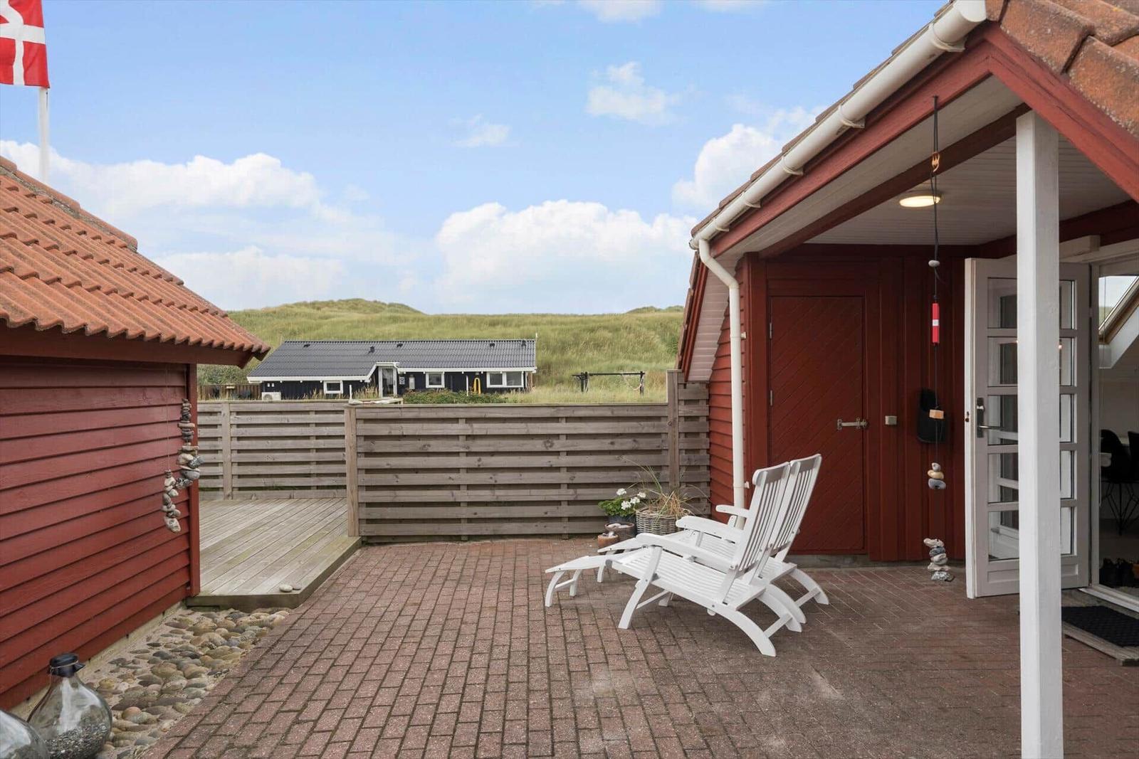 Terrasse mit weißen Liegestühlen, rotem Holzhaus und Blick auf grüne Dünen.