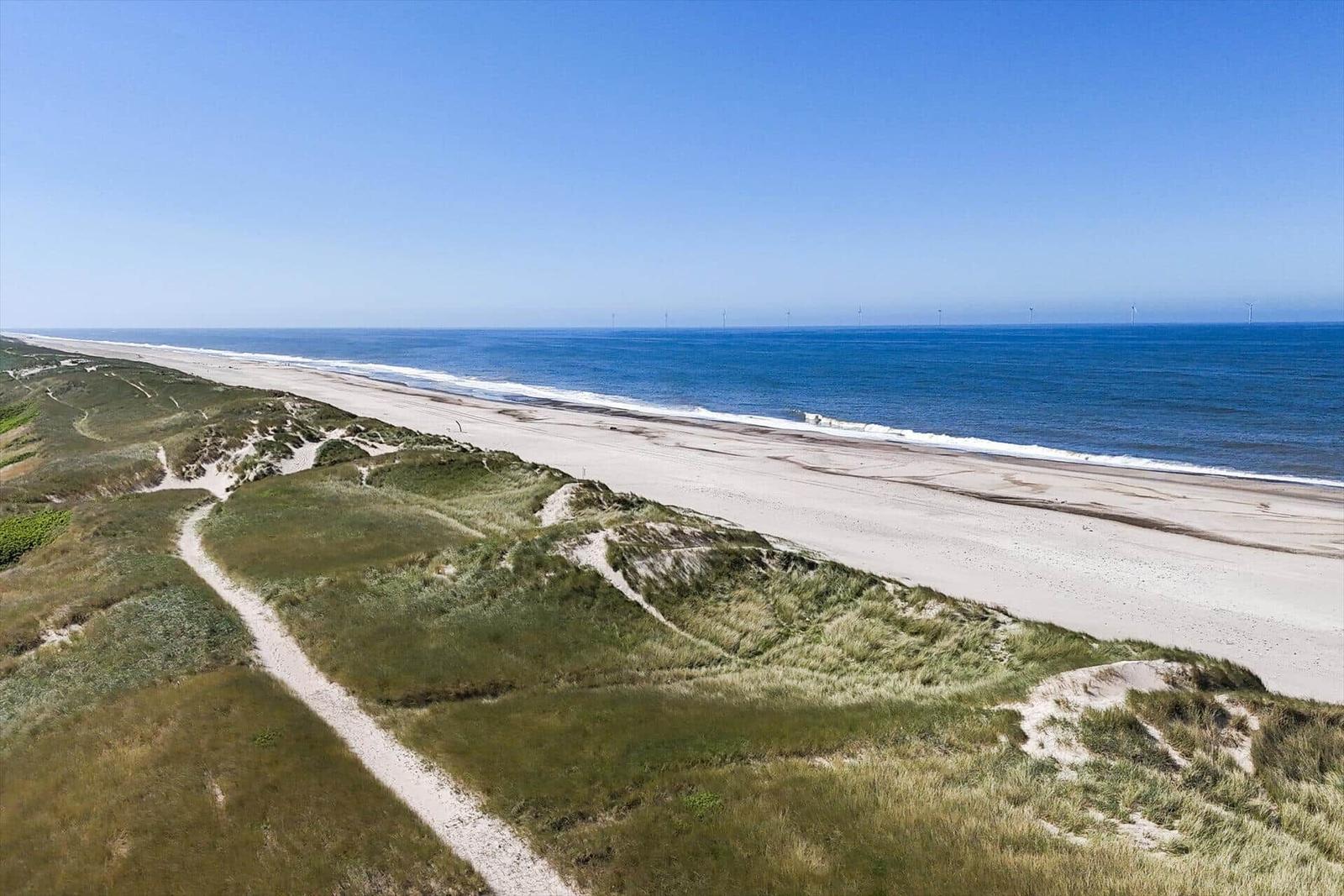 Strand mit Dünen und Windkraftanlagen im Hintergrund.