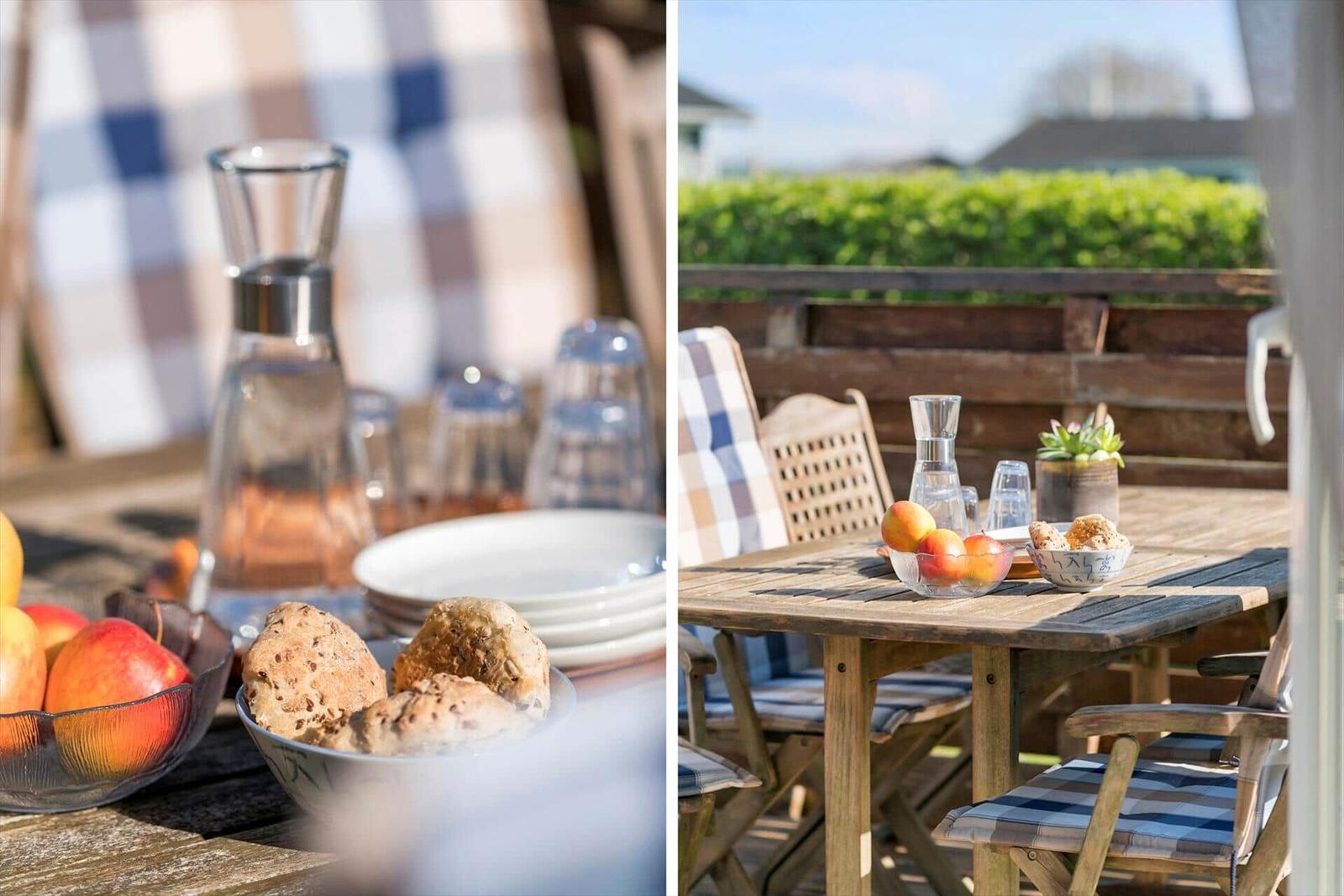 Wooden table with fruit and bread on terrace with chairs and hedge in background.