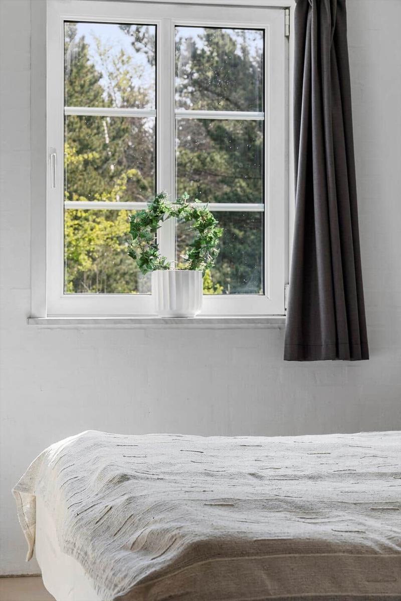 Ein Bett mit einer hellen Bettdecke. Ein Fenster mit grüner Pflanze und grauen Vorhängen.