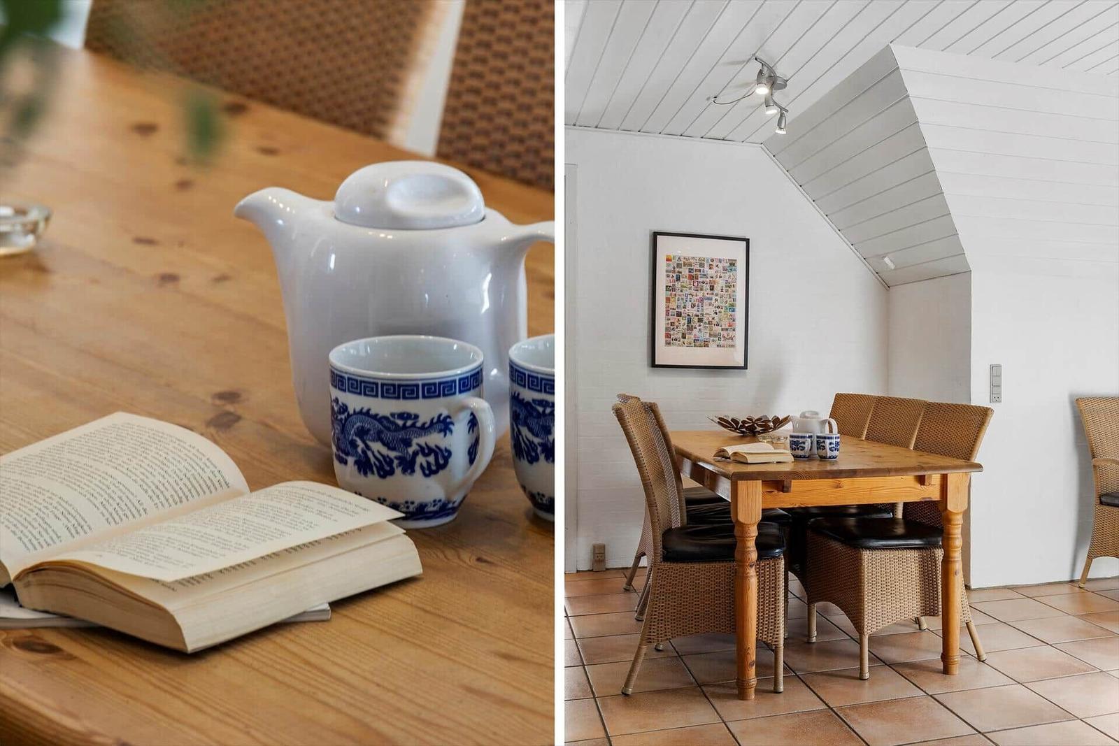 Holztisch mit Teekanne und Buch in einem Raum mit schrägem Dach und Wickerstühlen.