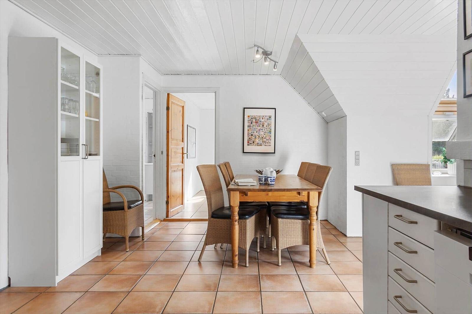 Esszimmer mit Holztisch, Stühlen und Küchenzeile. Weiße Wände und Holzdecke.