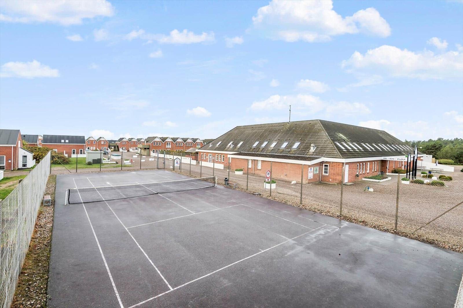 Tennisplatz mit Umzäunung und angrenzenden Gebäuden unter blauem Himmel.