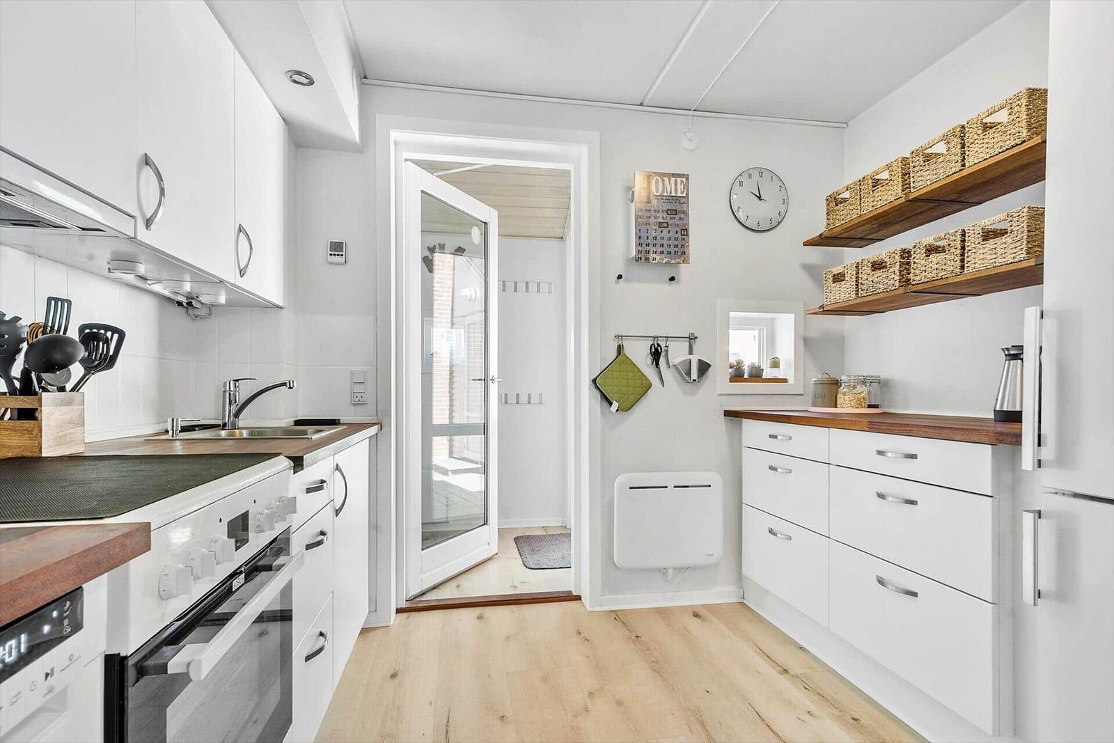 Küche mit weißen Schränken, Holzoberflächen und integriertem Ofen. An der Wand hängen Körbe und ein Wanduhr.