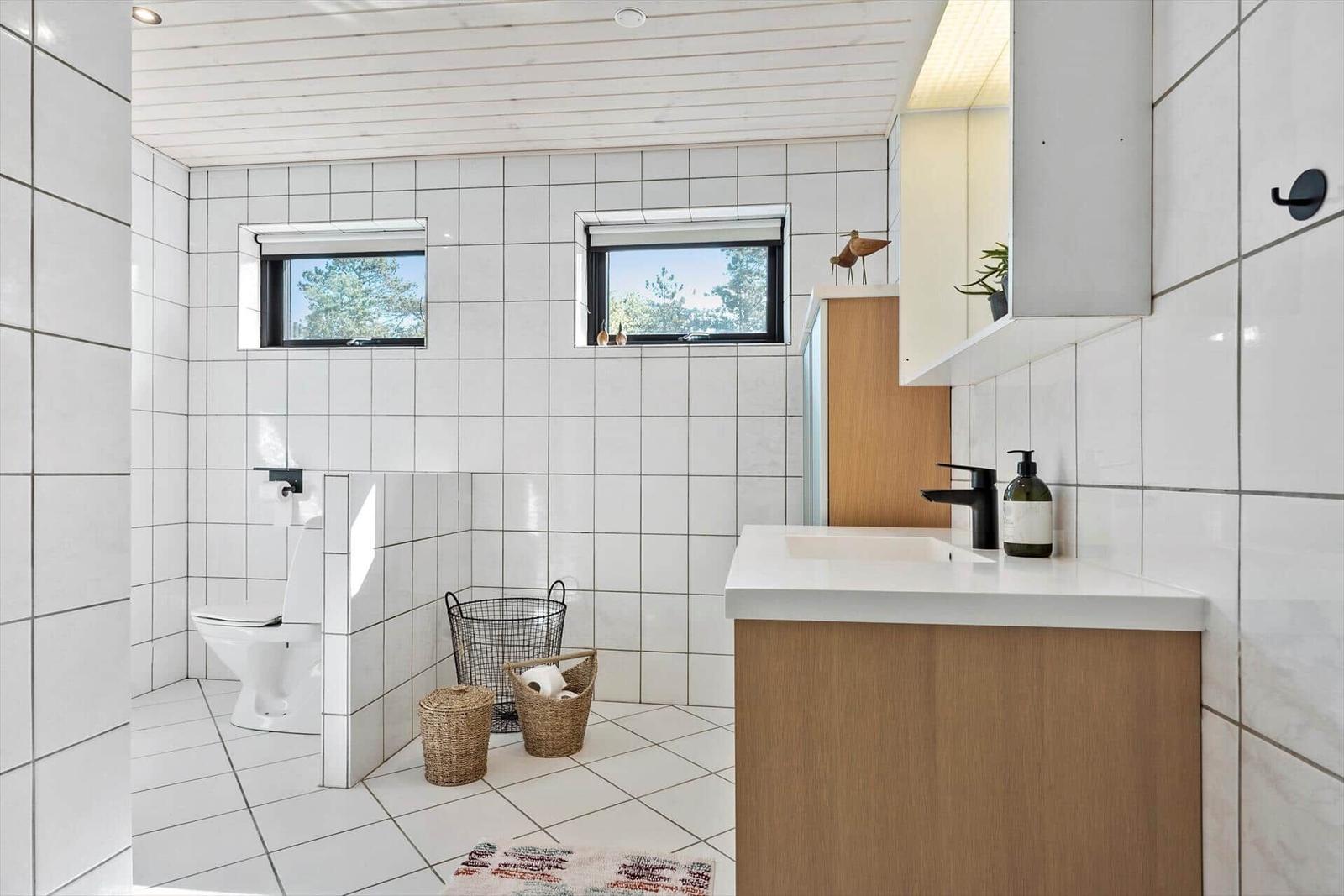 Badezimmer mit weißen Fliesen, zwei Fenstern und Holzwaschtisch.
