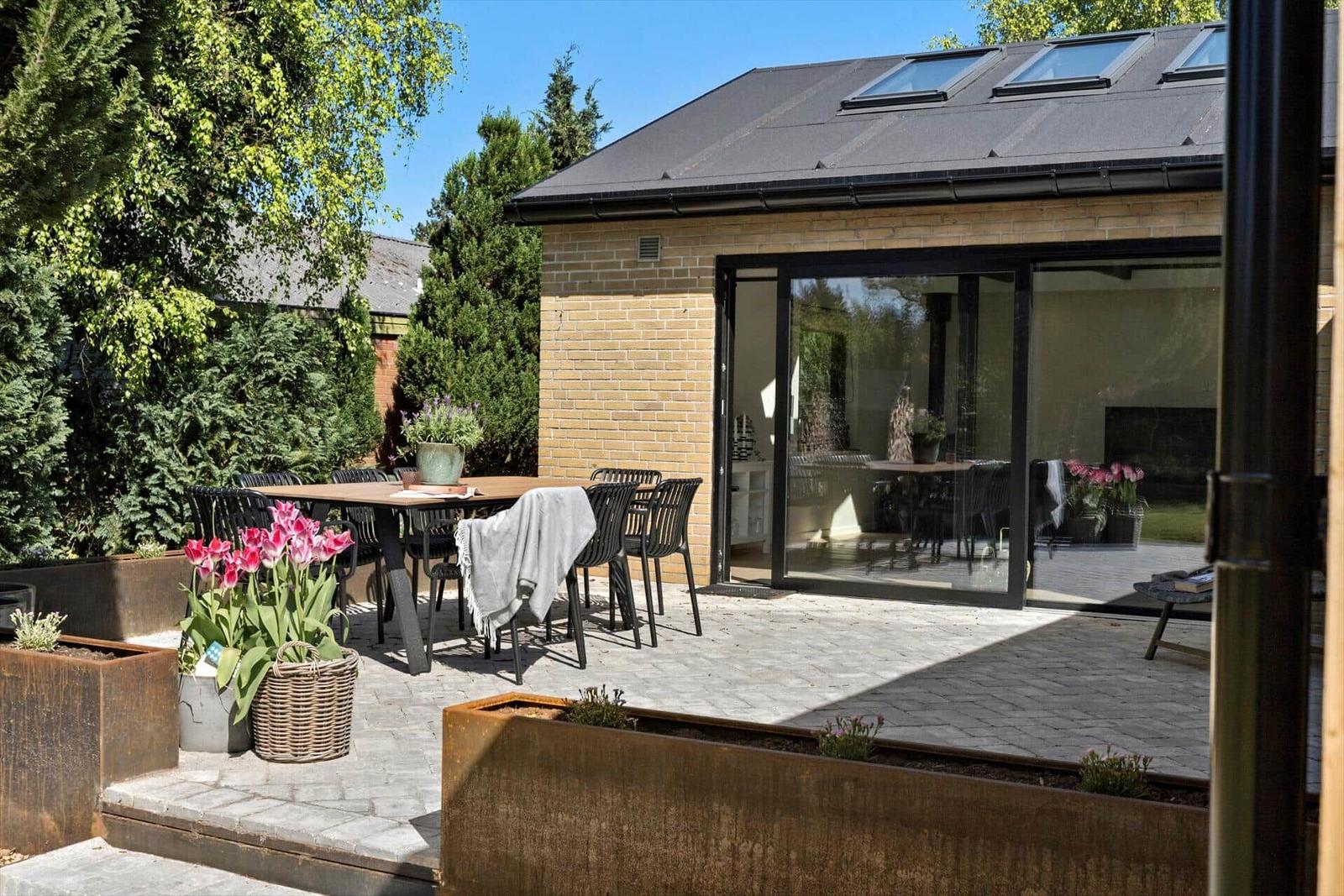 Terrasse mit Tisch, Stühlen und Blumen. Glasfront führt ins Haus. Hintergrund mit Bäumen.