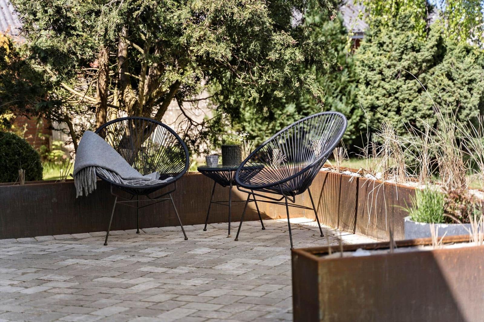 Terrasse mit zwei schwarzen Stühlen und Tisch, umgeben von Bäumen und Pflanzen.