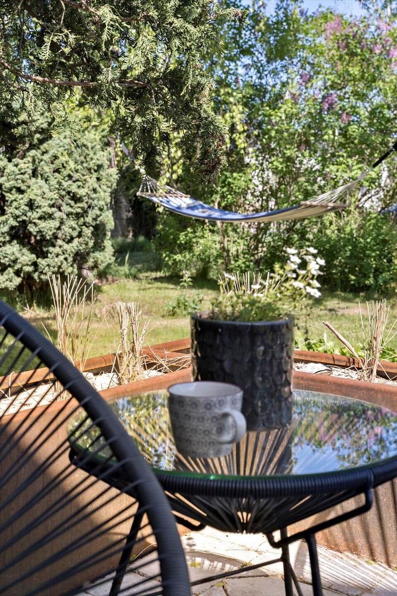 Garten mit Hängemattensitz, Tisch und Tasse. Grünflächen und Bäume im Hintergrund.