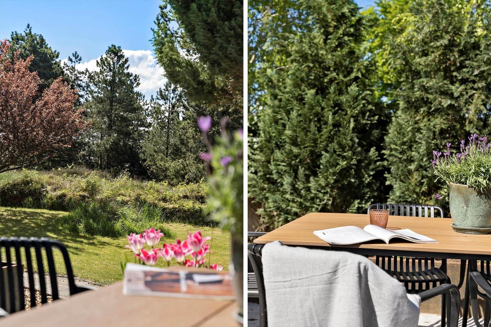 Terrasse mit Tisch, Stühlen und Blumen. Hintergrund: grüne Bäume und Wiese.