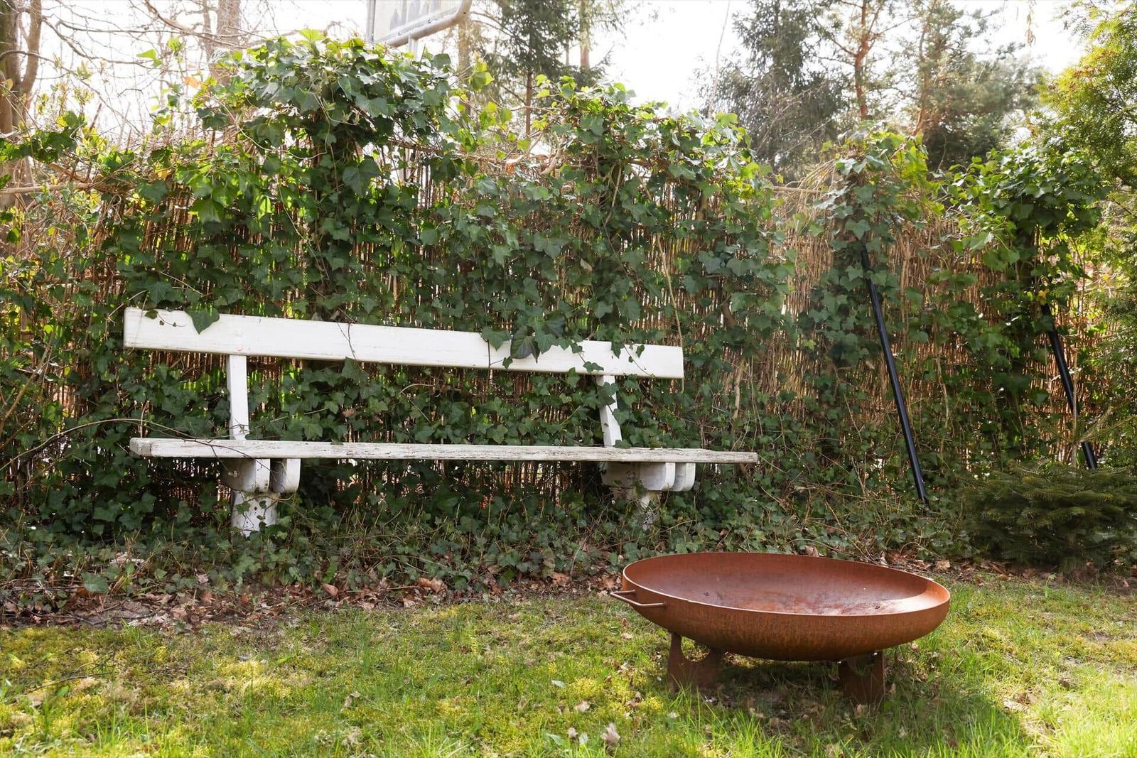 White bench and round fire pit in green garden with ivy on fence.