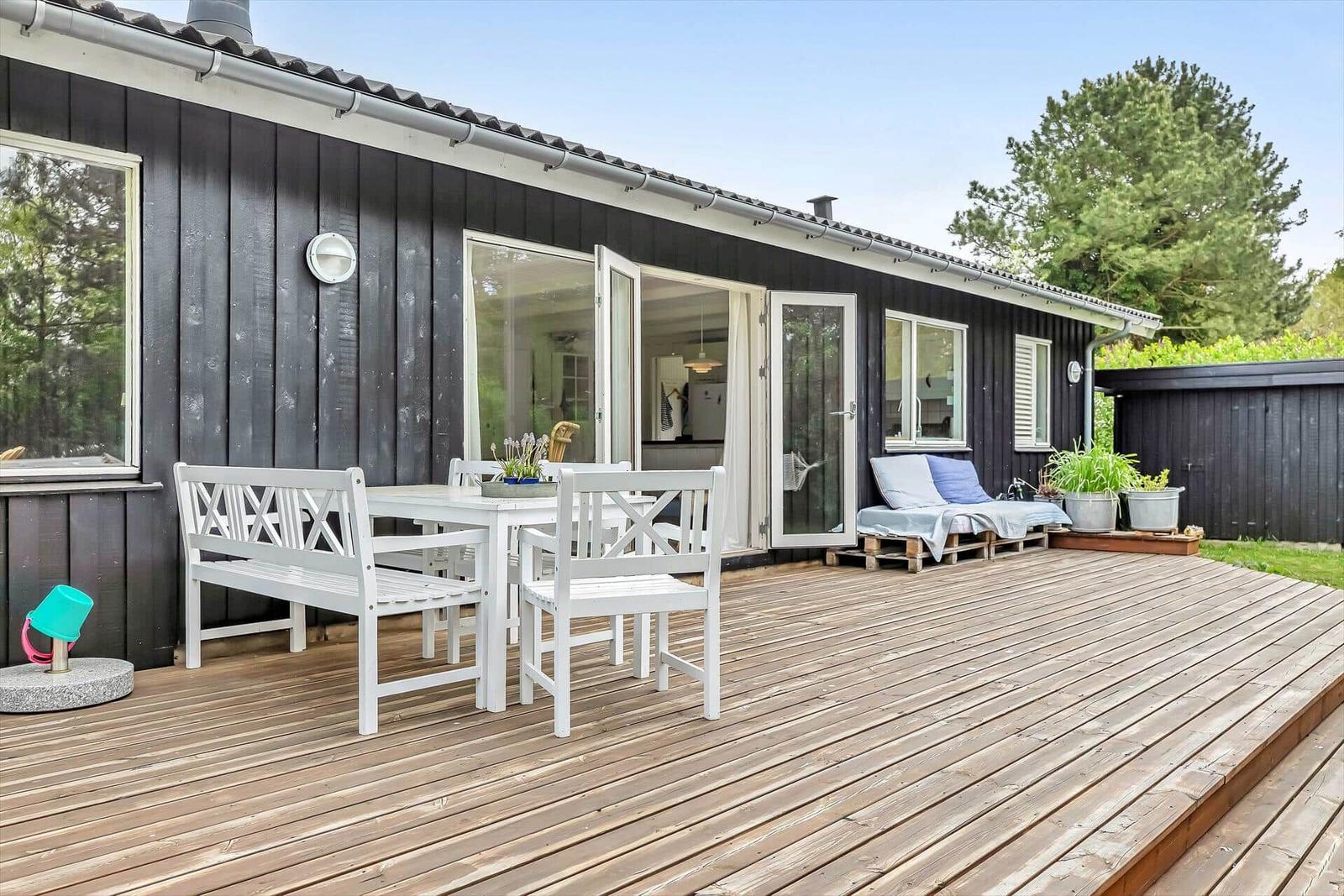 Terrasse mit weißem Tisch und Stühlen, Sitzbank und Pflanzen vor einem Haus mit dunkler Fassade.