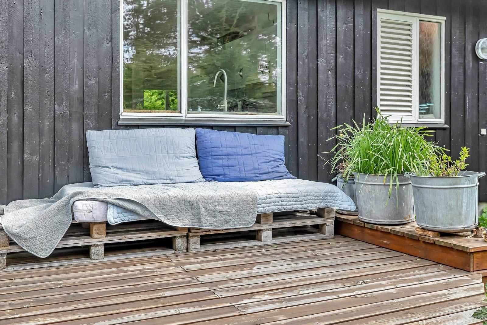 Außenliegende Matratze auf Paletten mit Kissen und Decke vor dunklem Holzhaus.
