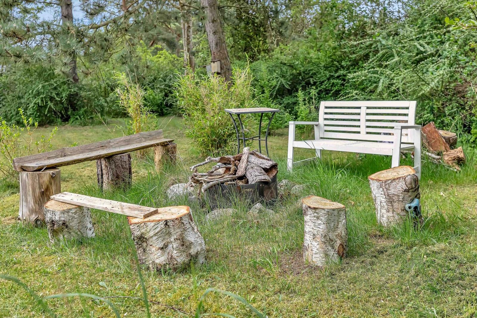 Außenbereich mit Holzbank, weiße Bank und Feuerstelle im Gras.
